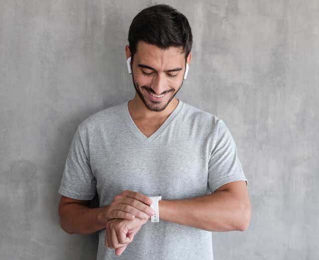 3Young-handsome-man-wearing-wireless-headphones-and-t-shirt,-checking-smart-watches-with-touch-screen,-standing-against-gray-textured-wall-with-copy-space-1018067034_2125x1417